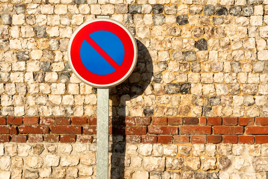 A circular no parking traffic sign fixed to a dark metal bracket mounted on a red brick wall exterior of a building. The sign features a blue background with a red border and a central red slash indic