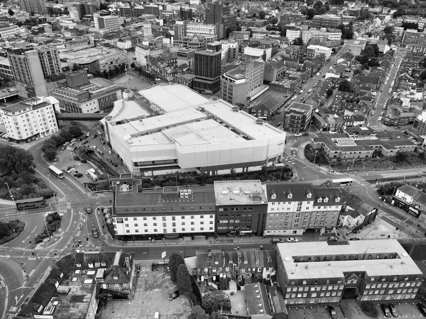 An aerial black-and-white photograph of a busy urban area showing a multi-story office building and residential apartments surrounding a network of roads and roundabouts. On the pavement near the buil