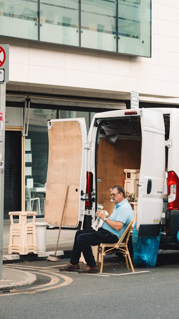 A man seated on a wooden chair outside a commercial building with large glass windows, unloading or preparing furniture and packing materials from a white cargo van parked on the street. The van's rea