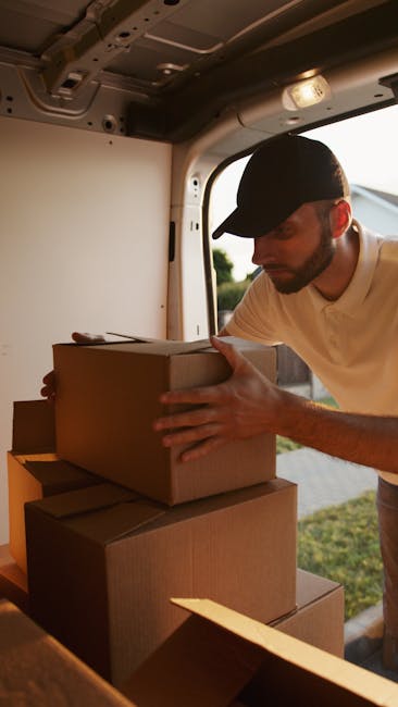 A man seated on a wooden chair outside a commercial building with large glass windows, unloading or preparing furniture and packing materials from a white cargo van parked on the street. The van's rea